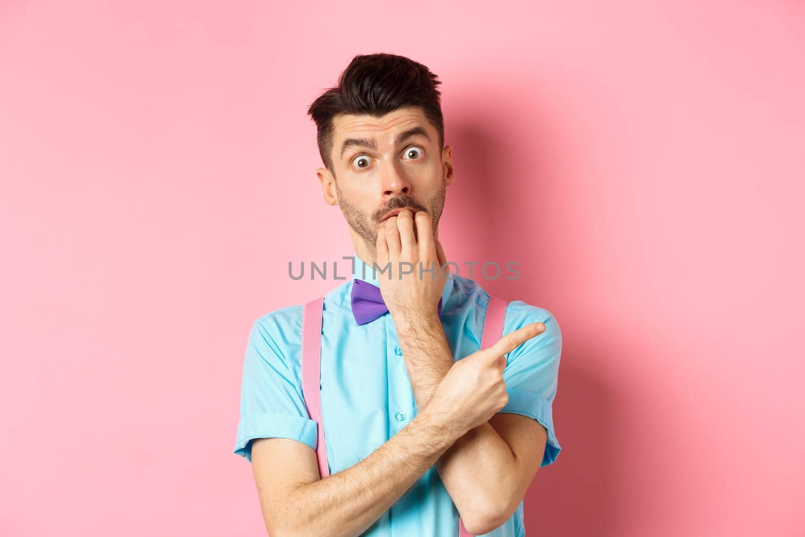 Intrigued guy in bow-tie biting fingernails from temptation, pointing fingers right at something he wants, standing over pink background.