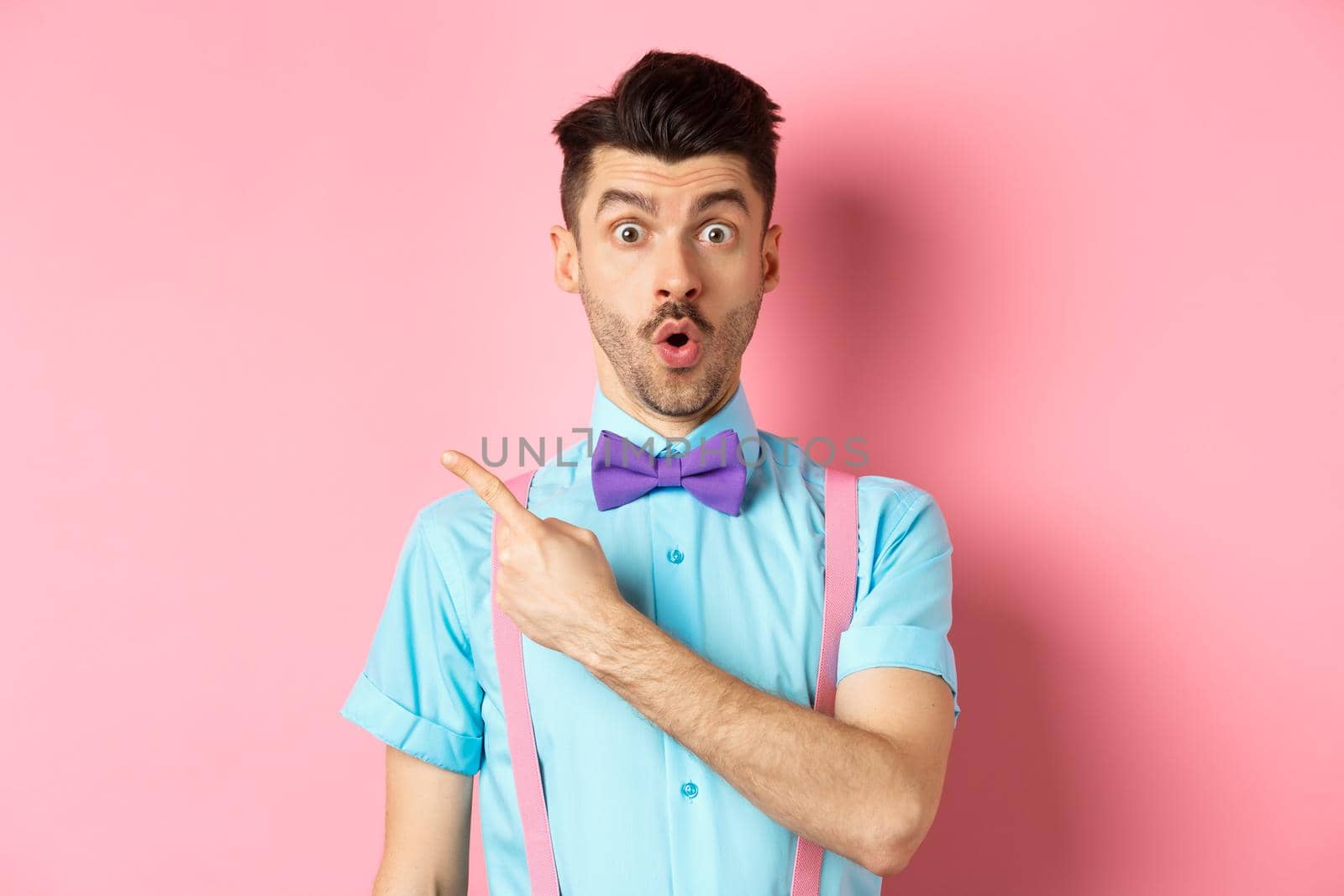 Check it out. Amazed caucasian man with moustache, wearing bow-tie, saying wow and pointing left, showing advertisement, standing on pink background.
