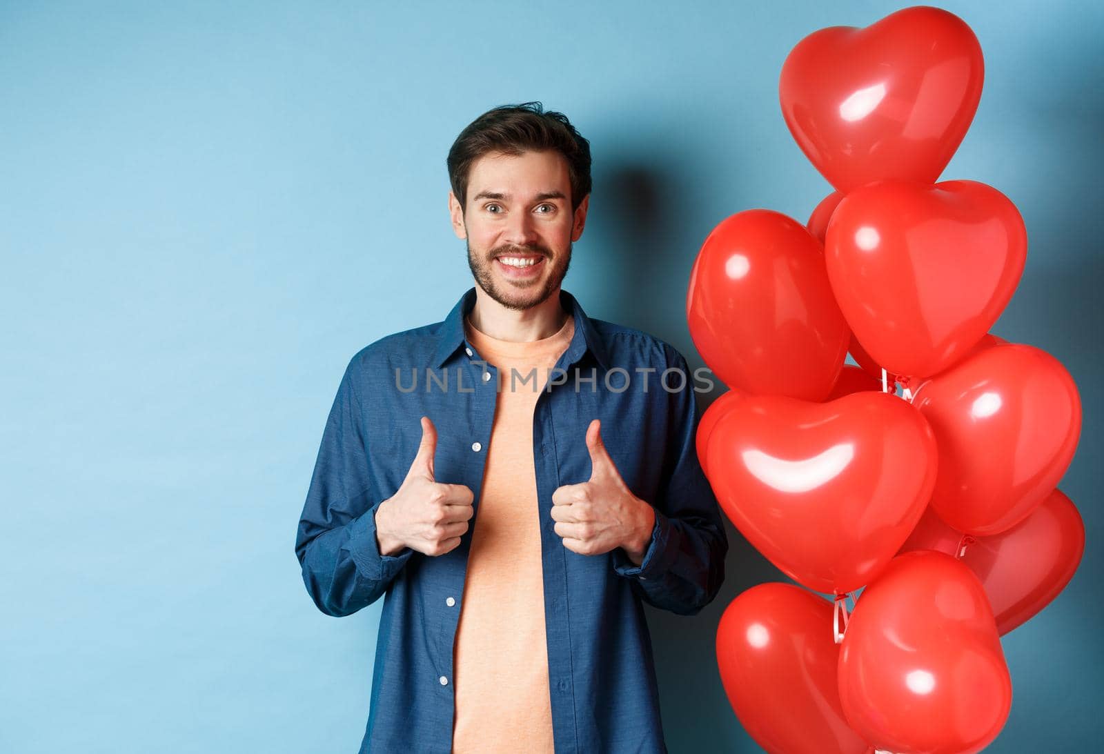 Happy valentines day. Cheerful boyfriend showing thumbs up in approval, standing with red hearts balloons for lover, blue background.