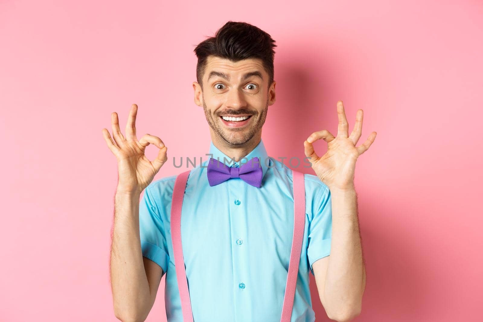 Cheerful handsome man smiling, showing okay gestures in approval, praising good choice, liking advertisement, standing on pink background.