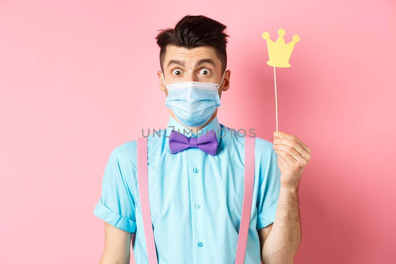 Covid, pandemic and quarantine concept. Cute and silly guy in medical mask looking surprised, holding small crown for party, standing over pink background.