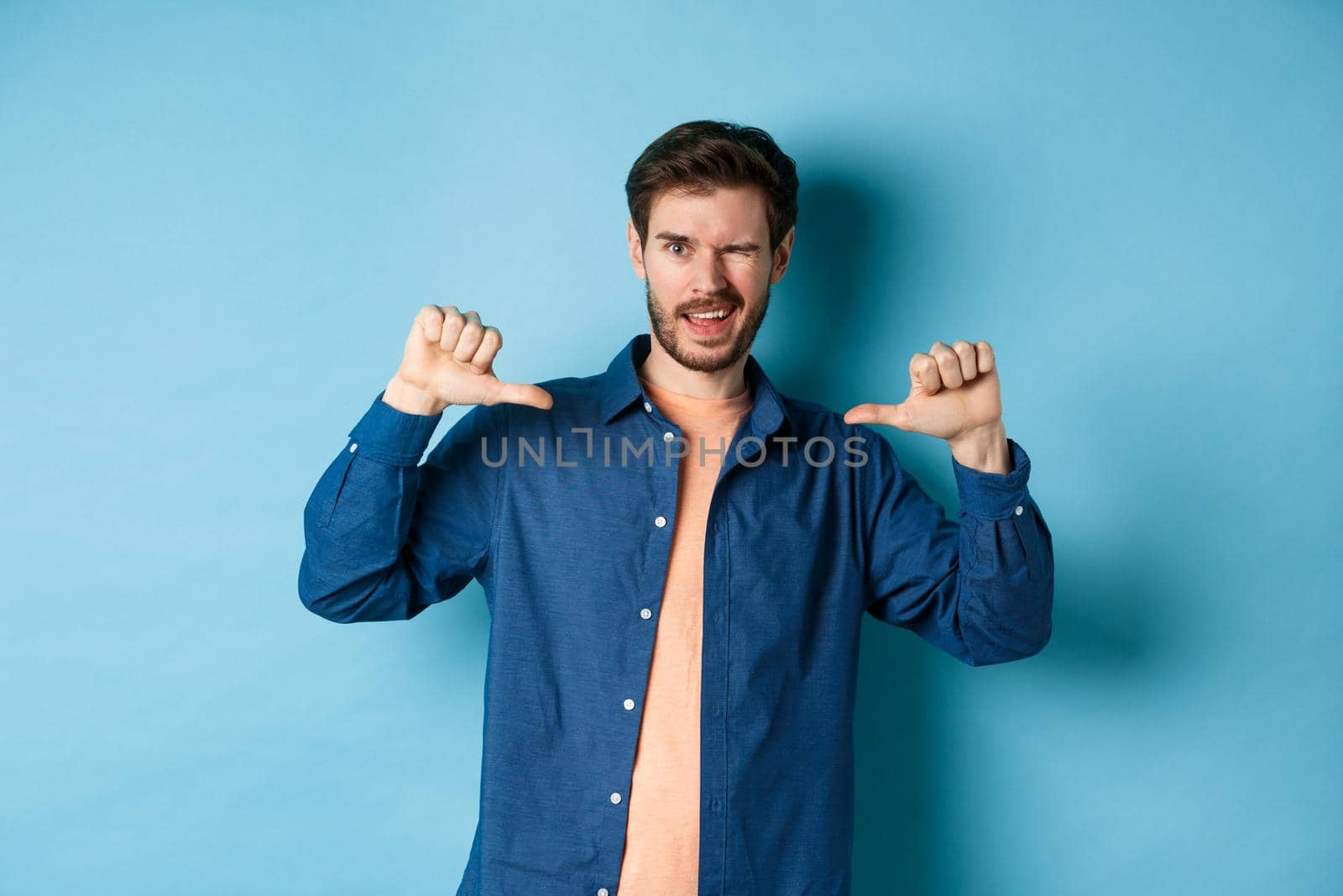 Confident man winking and pointing at himself, self-promoting, standing on blue background by Benzoix