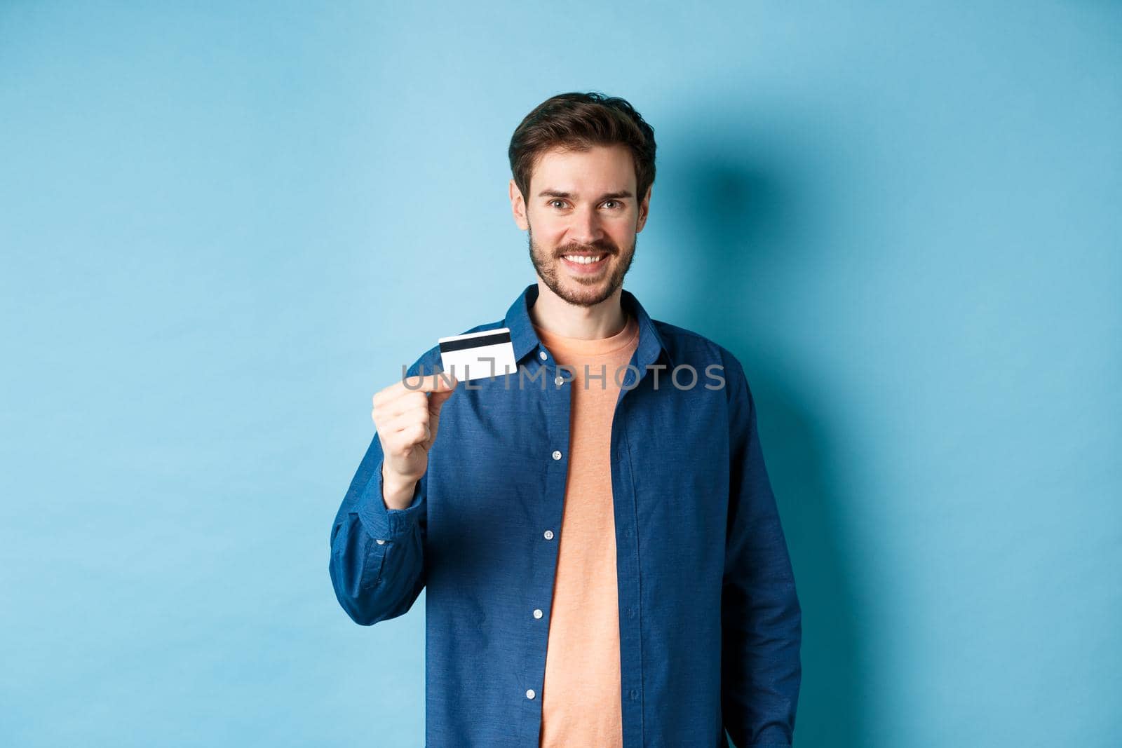Handsome smiling guy showing plastic credit card and looking satisfied, standing on blue background.