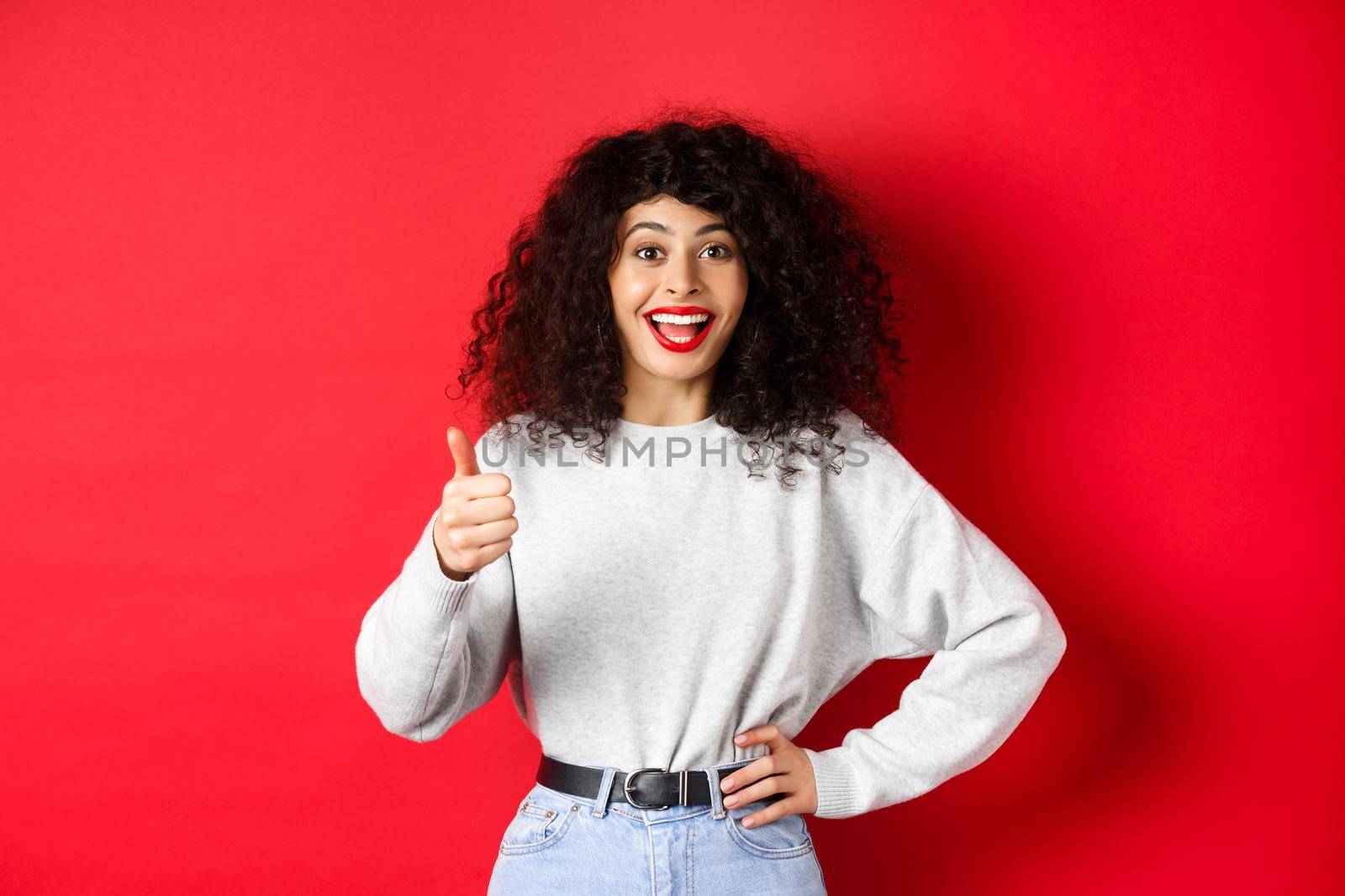 Happy young woman with curly hair praising good work, say well done and show thumb up gesture, approve and praise you, standing on red background.
