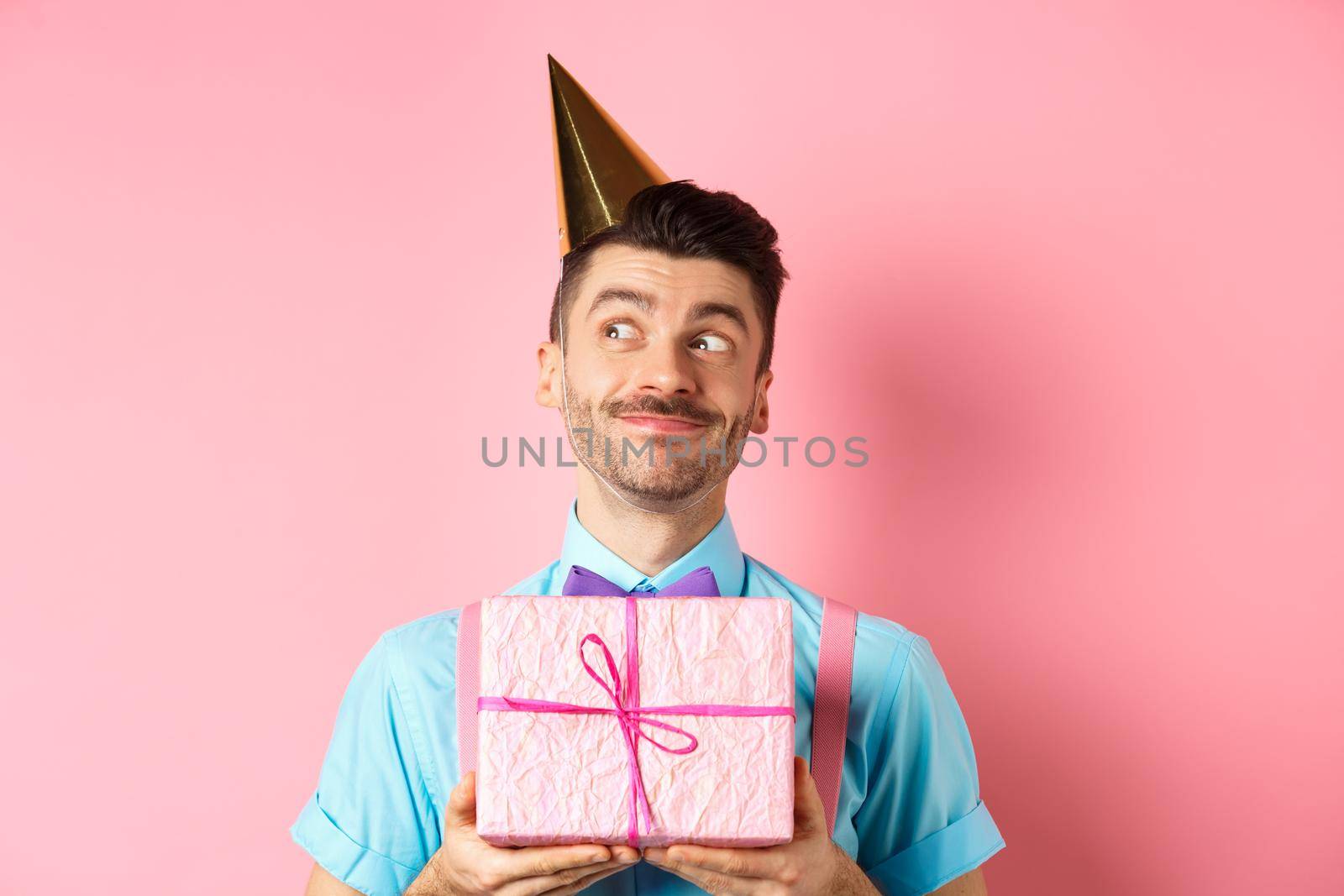 Holidays and celebration concept. Silly guy with moustache and bow-tie, wearing party hat, receive birthday gift and looking dreamy aside, standing over pink background.