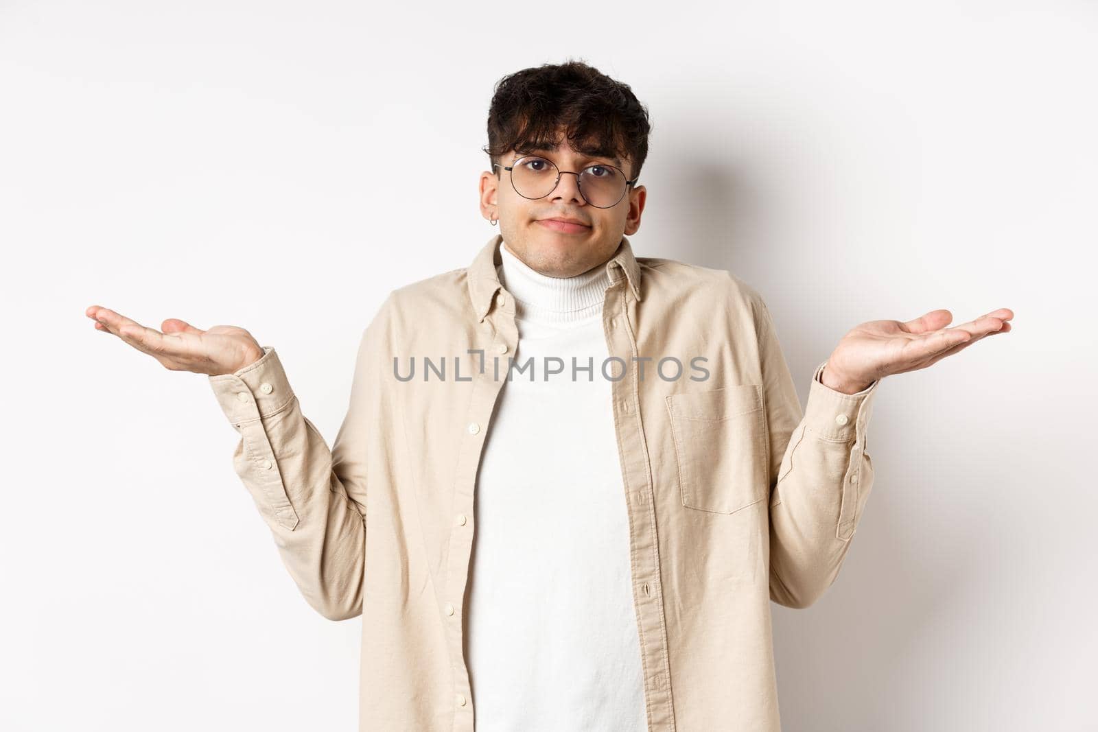Portrait of handsome young man looking confused, know nothing, shrugging shoulders and spread hands sideways clueless, standing on white background.
