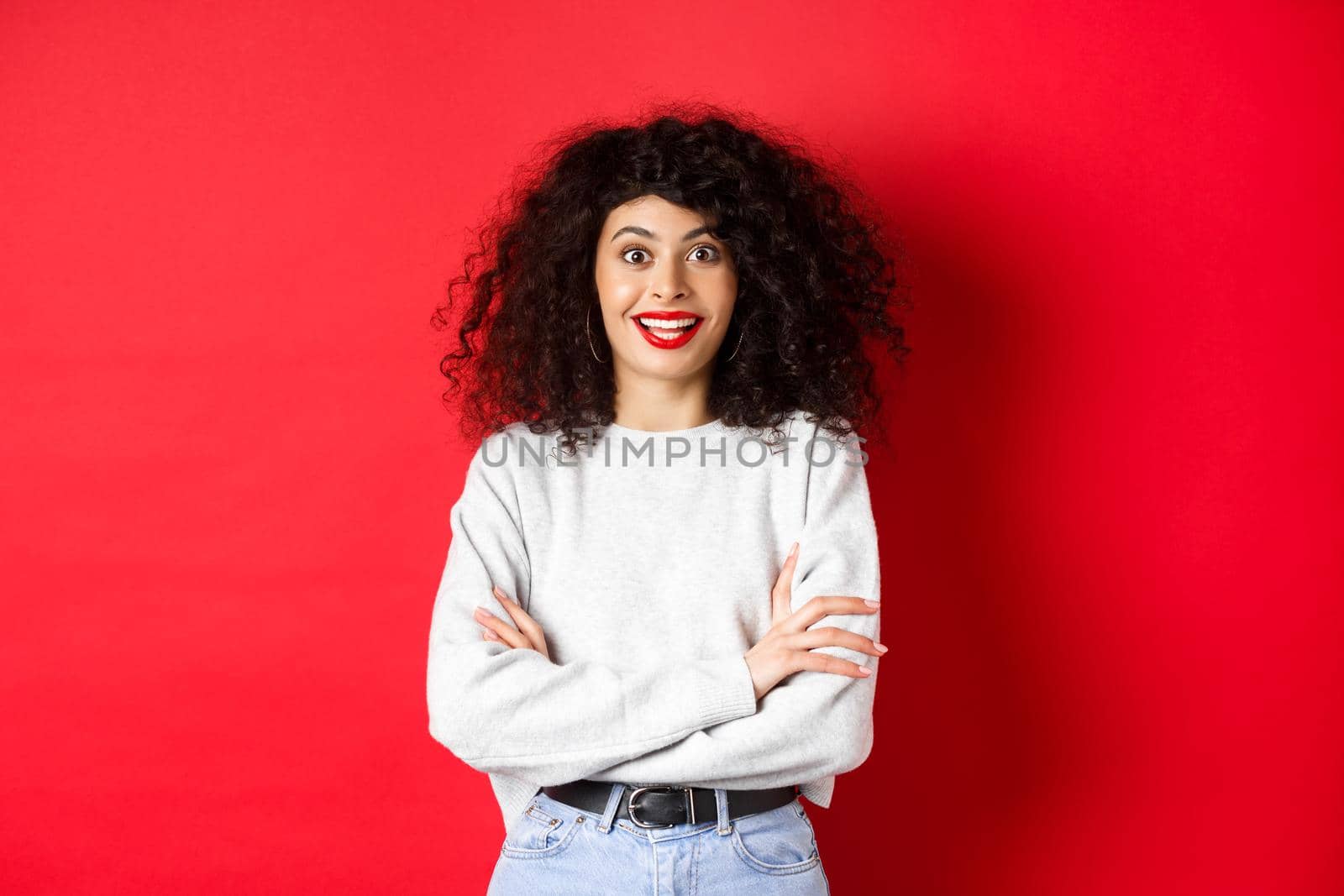 Cheerful young woman with curly hairstyle, raising eyebrows and looking surprised, hear interesting news, red background.
