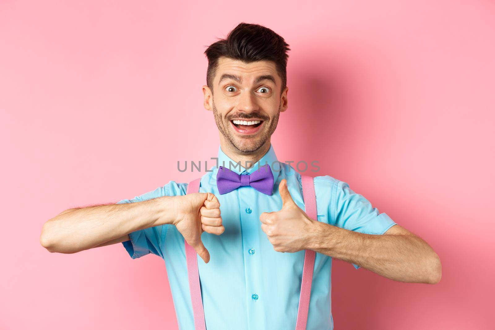 Funny young man smiling and showing thumbs up down, judging something and being indecisive, making choice, standing on pink background.