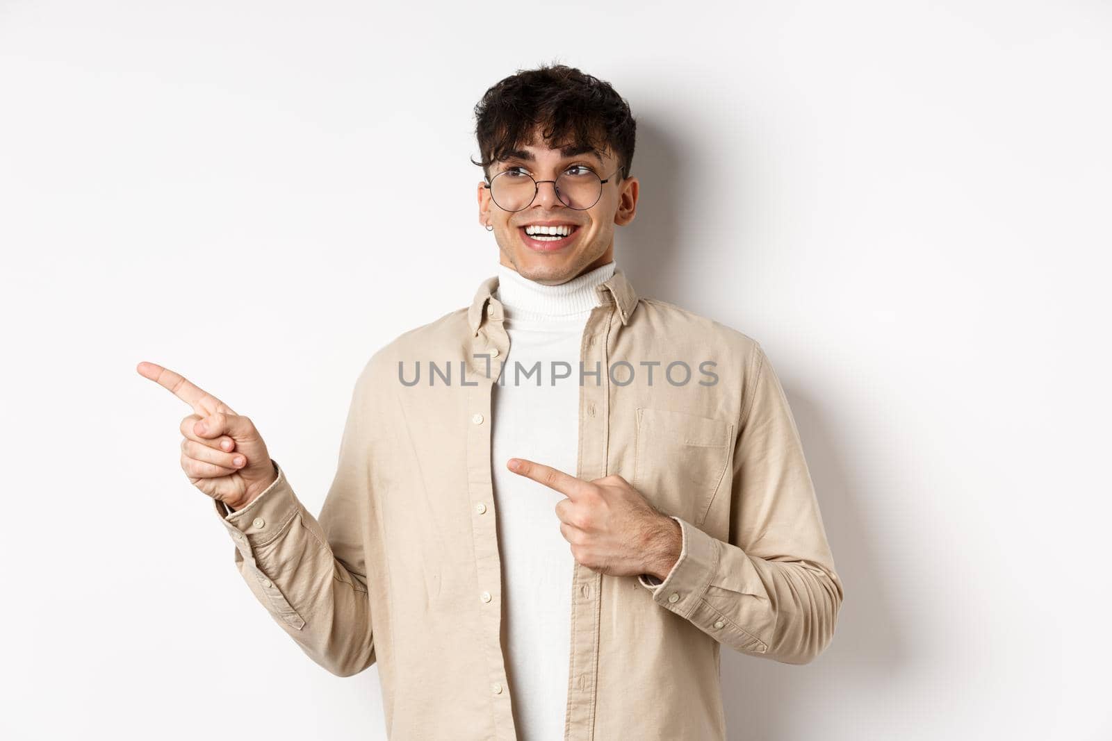 Excited and enthusiastic young man in glasses, pointing fingers and looking left with happy smile, standing in awe on white background by Benzoix