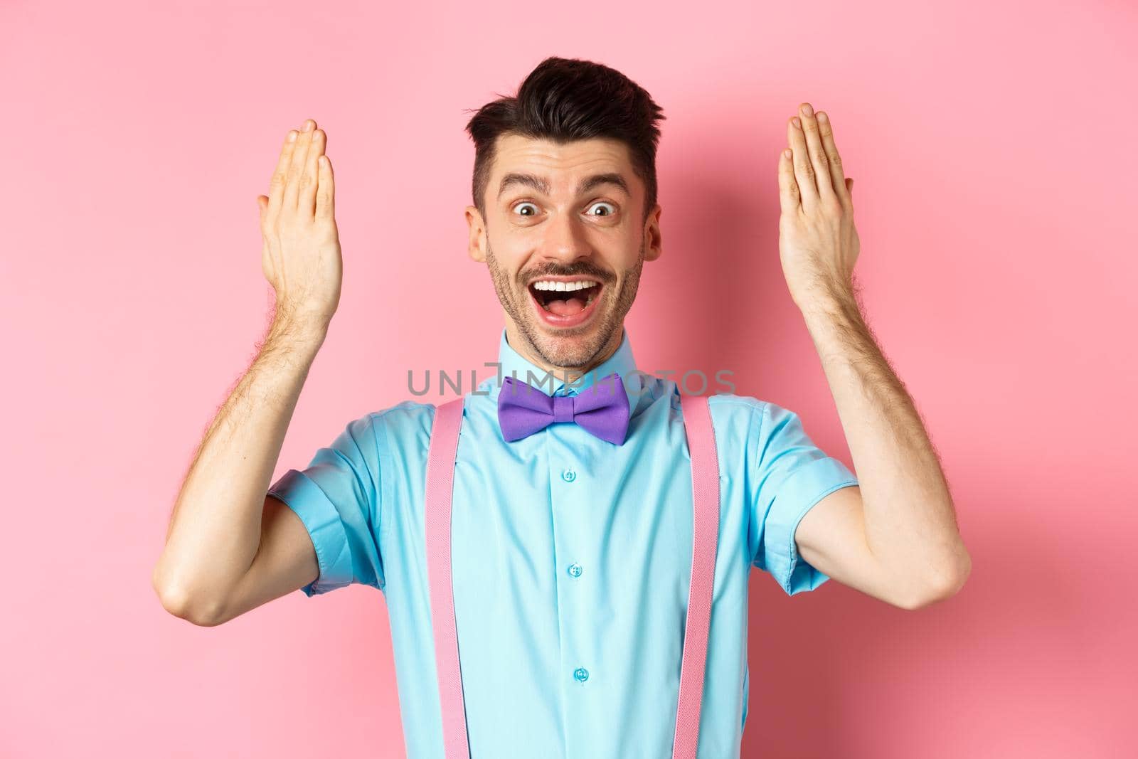 Cheerful young man open eyes for surprise present on holiday celebration, looking amazed at gift, standing on pink background.