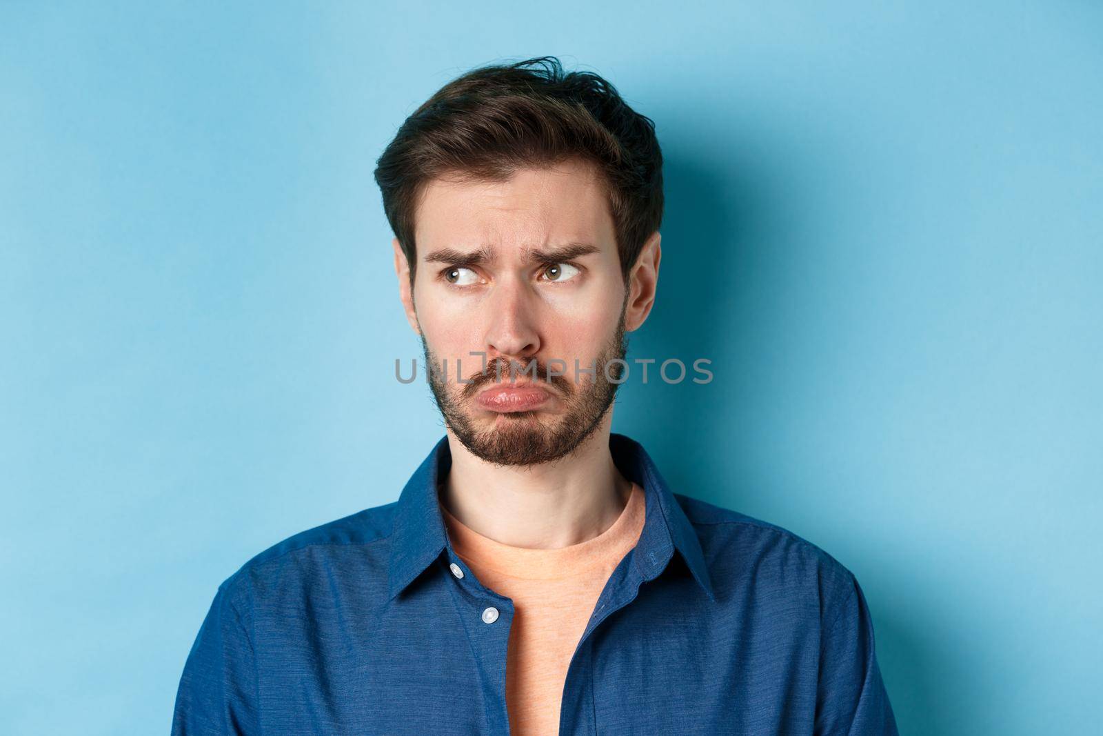 Miserable and disappointed guy acting childish, sulking and looking left at empty space with offended face, standing on blue background.