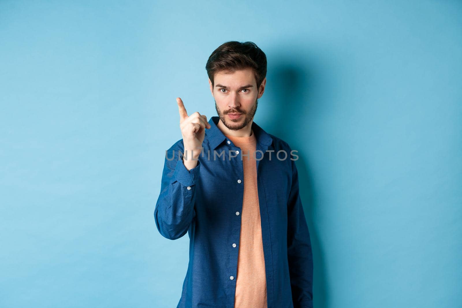 Serious young man scolding person, shaking finger disapprovingly, warn or prohibit something bad, standing on blue background.
