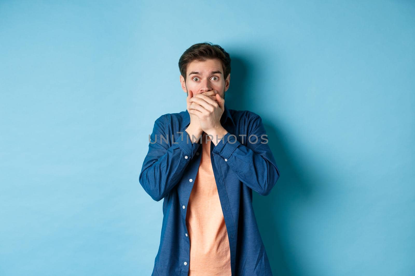 Shocked man covering mouth and gasping ambushed, witness something scary, standing on blue background.