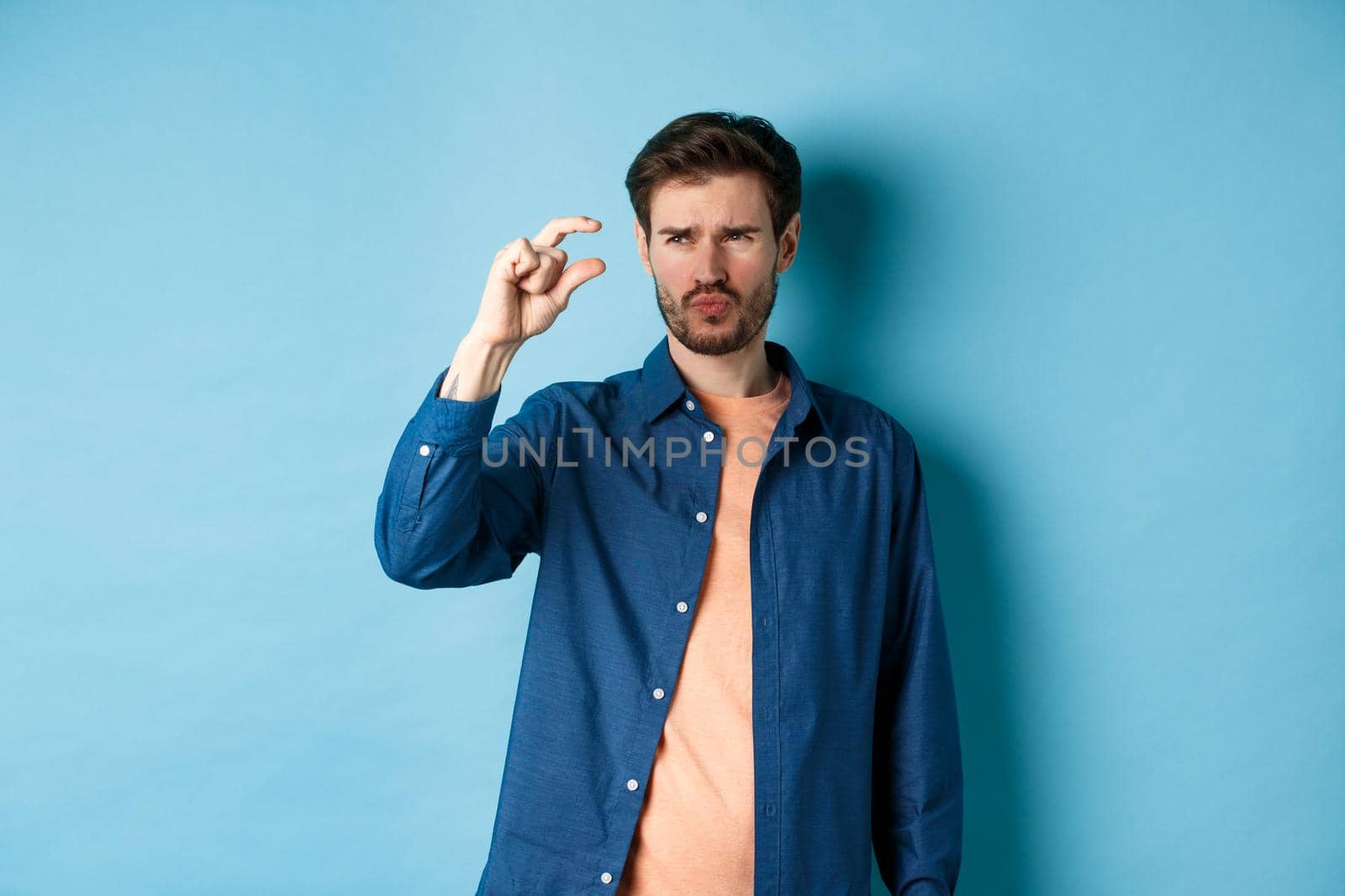 Confused guy showing small size and looking doubtful or skeptical, standing over blue background by Benzoix