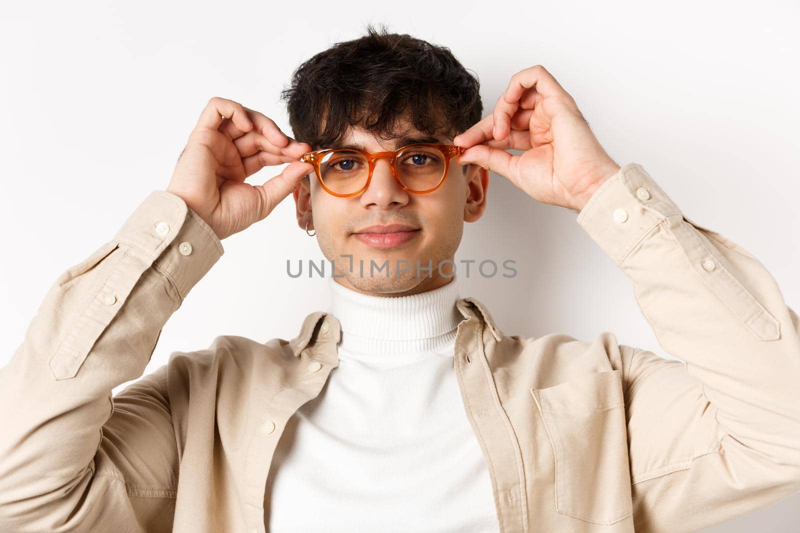 Close-up of stylish hipster guy trying eyewear at optician store, put on glasses and smiling, standing on white background by Benzoix