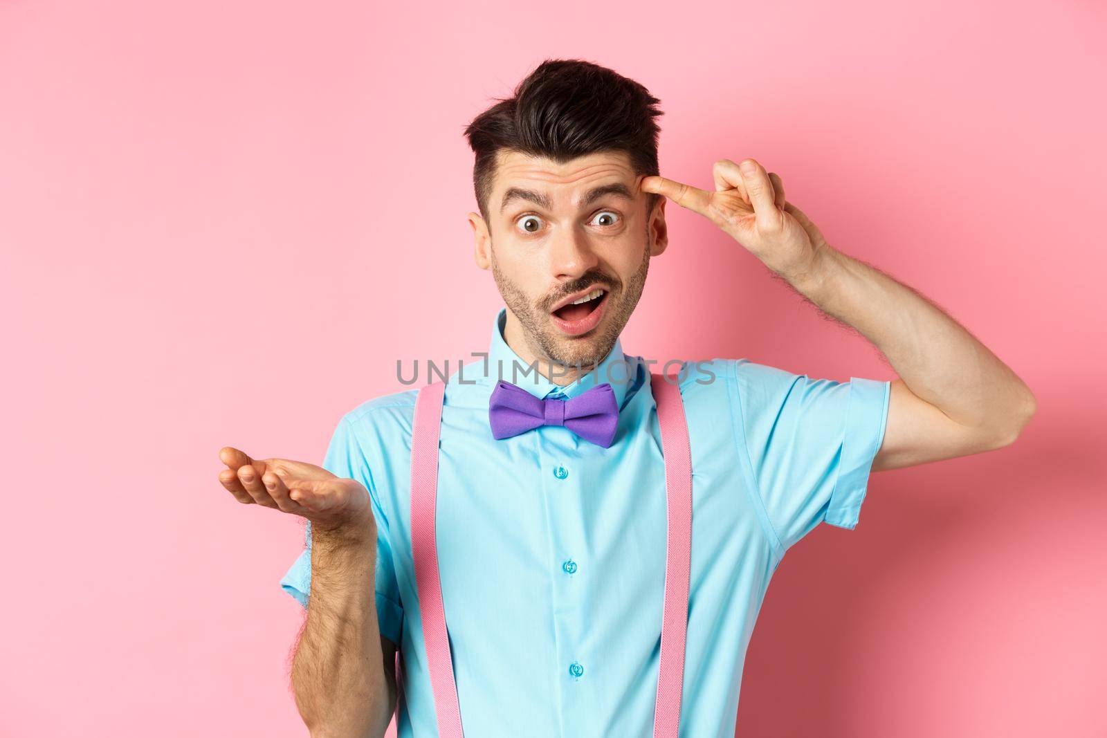 Annoyed guy mocking someone stupid, pointing finger at head and camera, are you stupid gesture, standing over pink background.