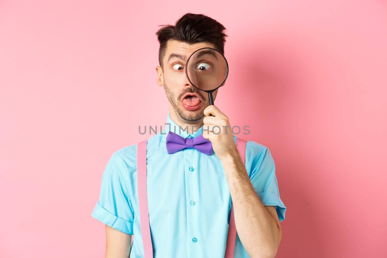 Funny man in bow-tie look through magnifying glass, squinting and making silly faces, standing on pink background.