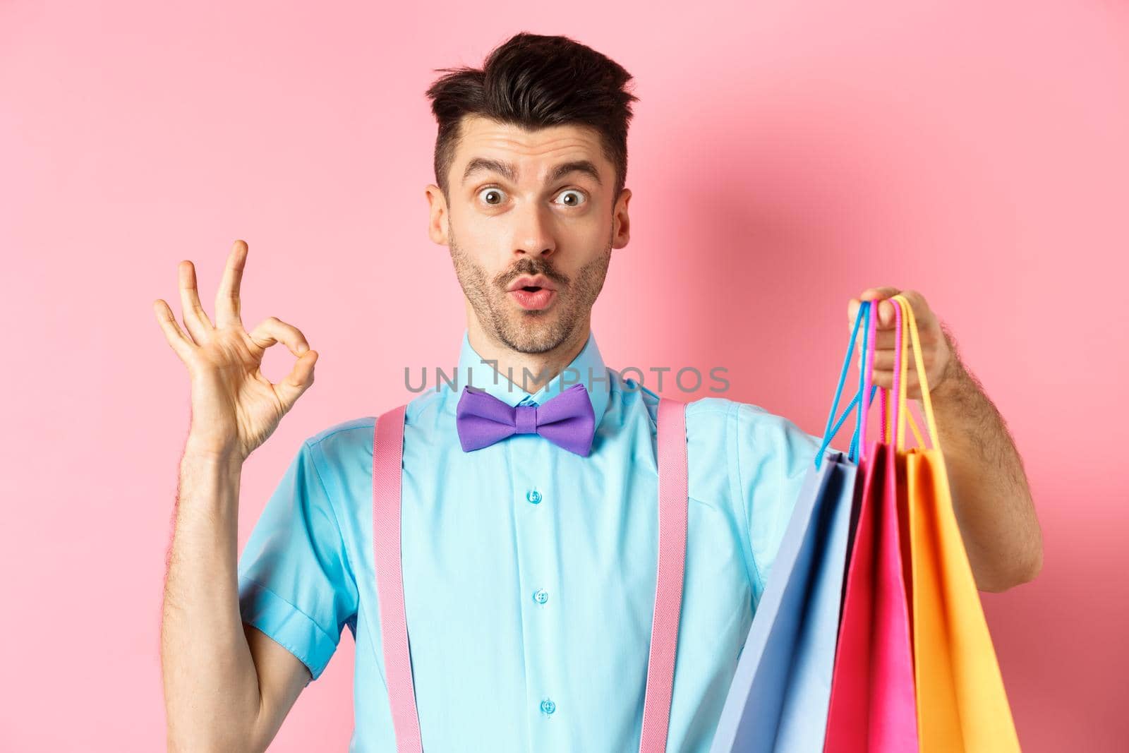Funny guy in bow-tie showing OK sign and shopping bags, praising good deal in shops, standing over pink background.