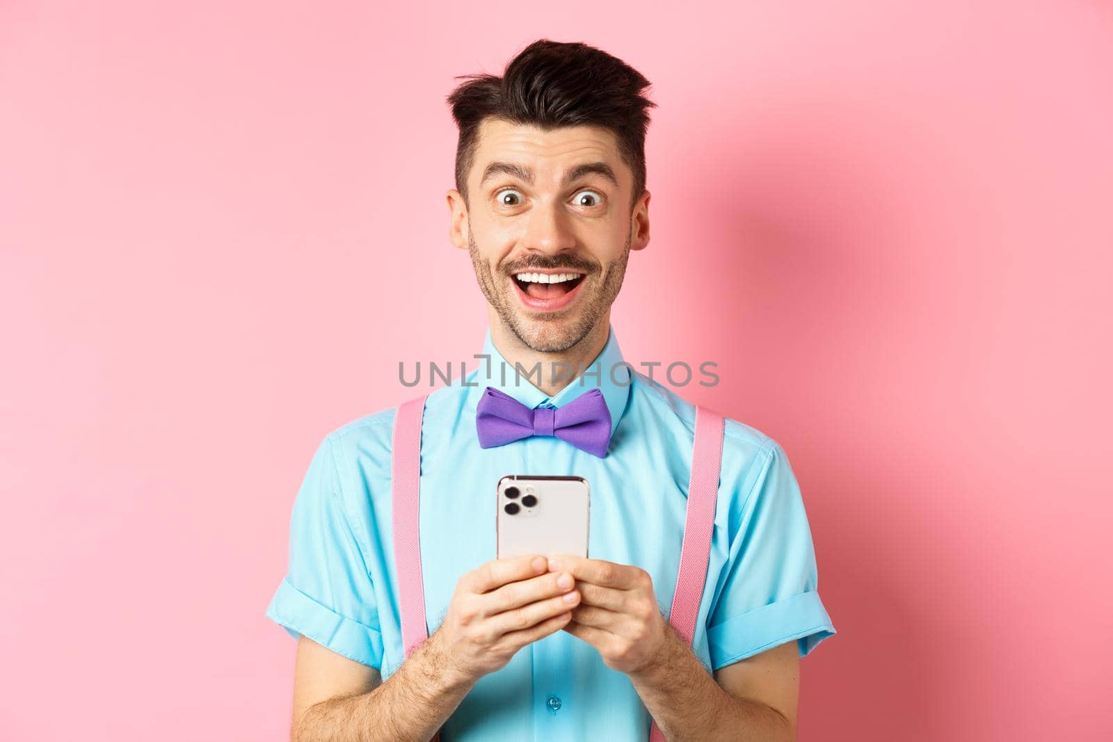 Online shopping. Happy man looking amazed after reading smartphone screen, smiling excited at camera, standing over pink background.