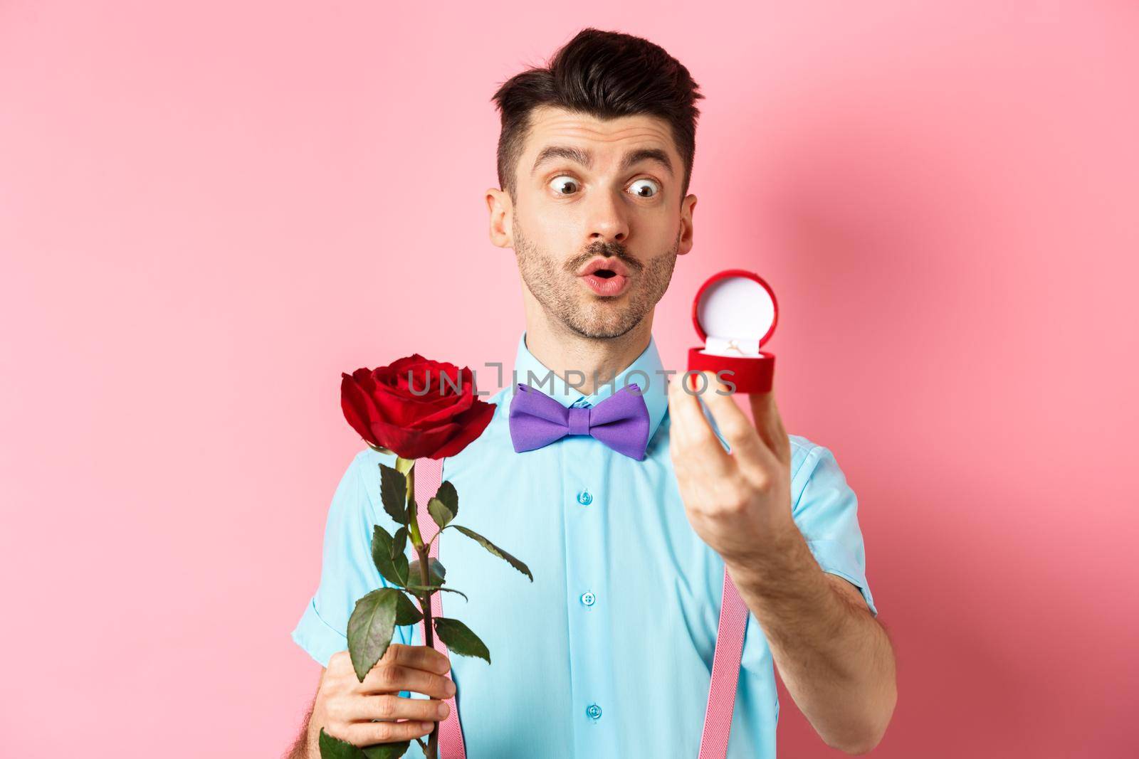 Valentines day. Romantic funny guy going to make wedding proposal, asking to marry him, holding red rose and looking at engagement ring with excitement, pink background.