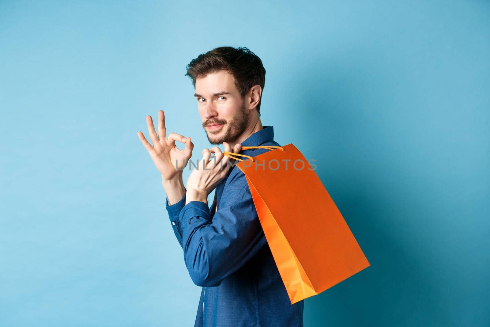 Handsome young man holding shopping bag on shoulder, turn around to look at camera and show okay gesture, recommending shop, blue background by Benzoix