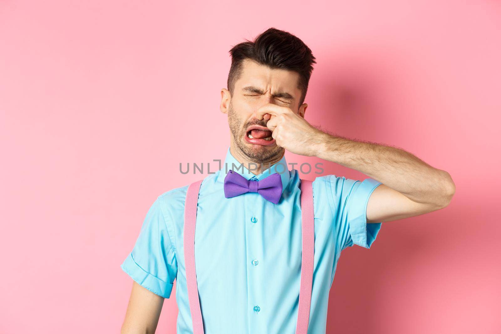 Image of man crying from disgusting smell, shut his nose from awful stink, standing in bow-tie and suspenders on pink background.
