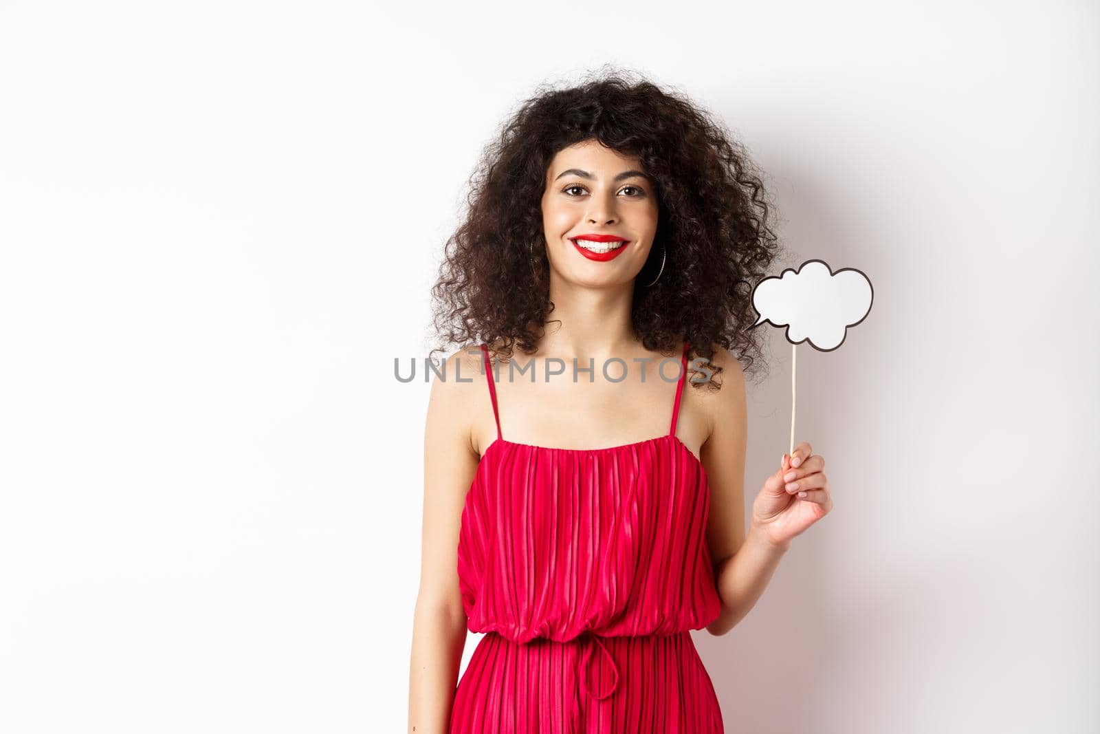 Happy stylish woman with curly hair, beauty makeup, holding comment cloud on stick and smiling, standing in red dress on white background.