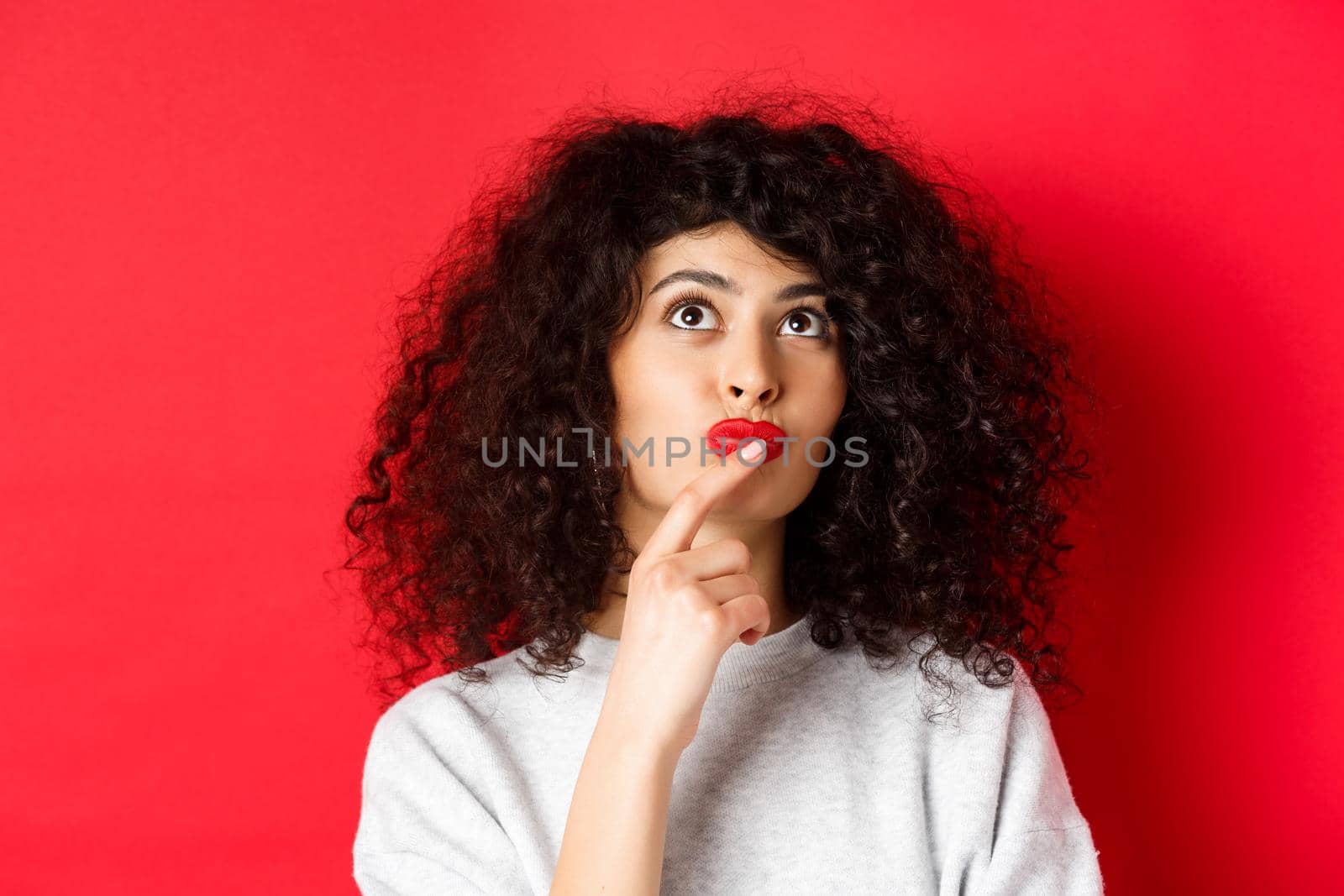 Close-up portrait of pensive young woman making choice, looking up thoughtful and serious, standing on red background.
