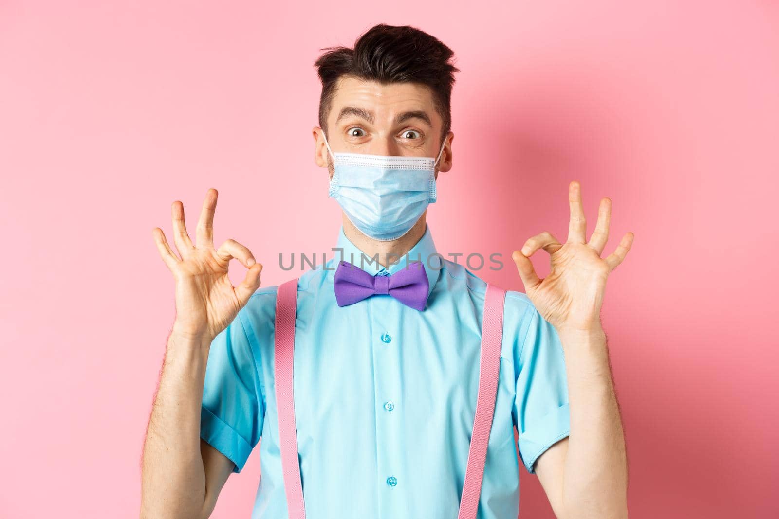 Coronavirus, healthcare and quarantine concept. Happy guy in medical mask and festive bow-tie showing all good gesture, make okay signs and smile at camera, pink background.