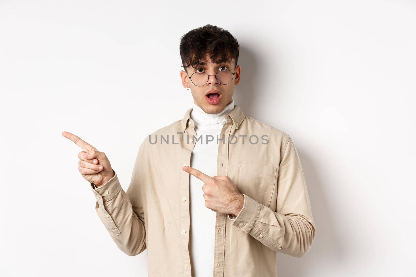 Portrait of young man in glasses drop jaw impressed, pointing fingers left at empty space with amazed face, standing on white background.