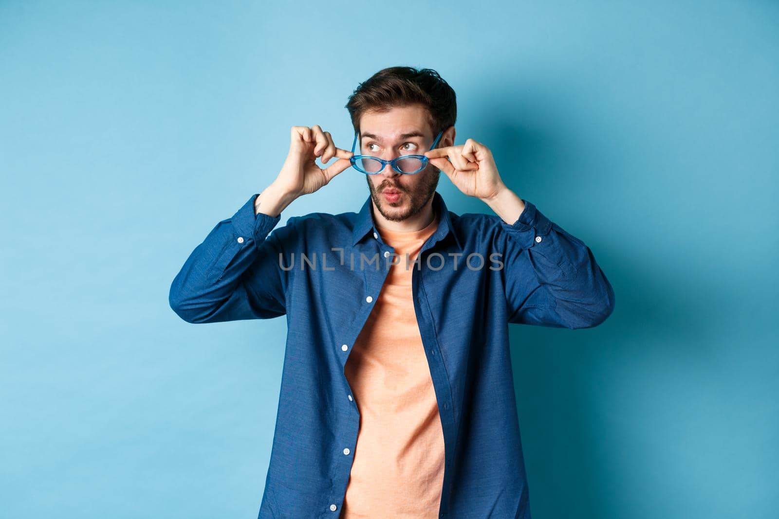 Image of caucasian guy takes-off sunglasses and looking aside with surprised face, checking out empty space for logo, standing on blue background.
