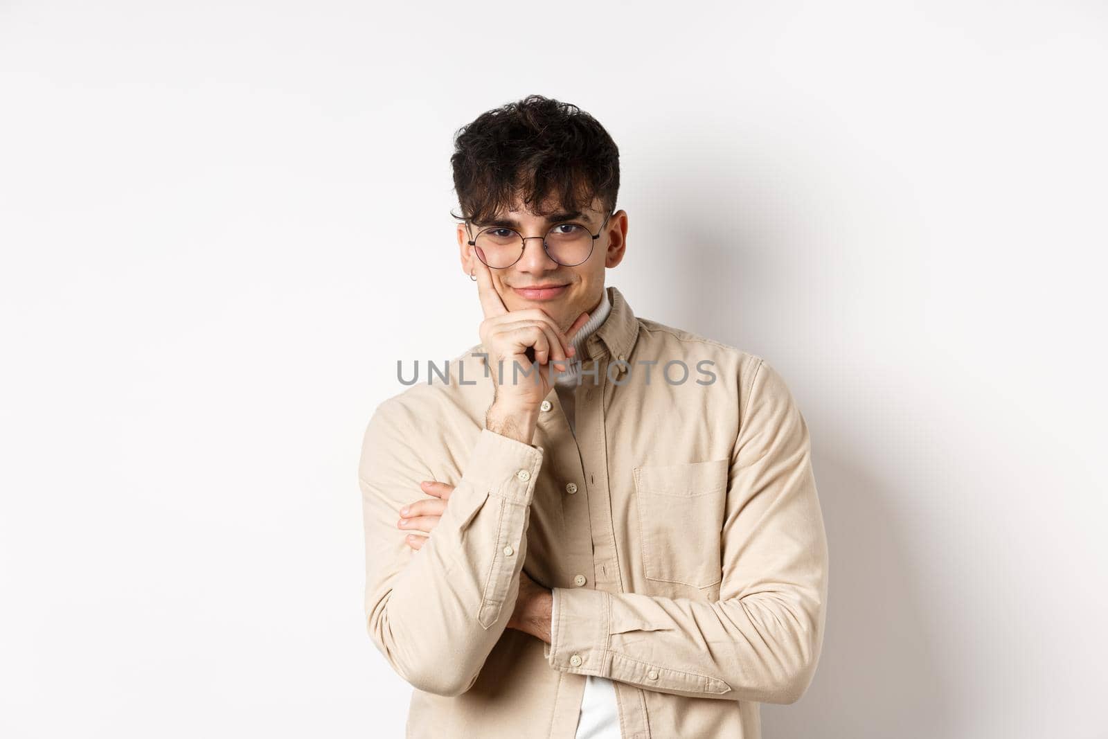 Cunning young man watching something interesting happen, lean face on arm and smiling devious, looking at camera intrigued, standing on white background.