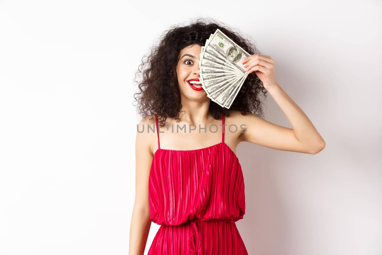 Happy curly woman winning money, holding dollar bills on half of face, smiling excited, standing in red dress on white background by Benzoix