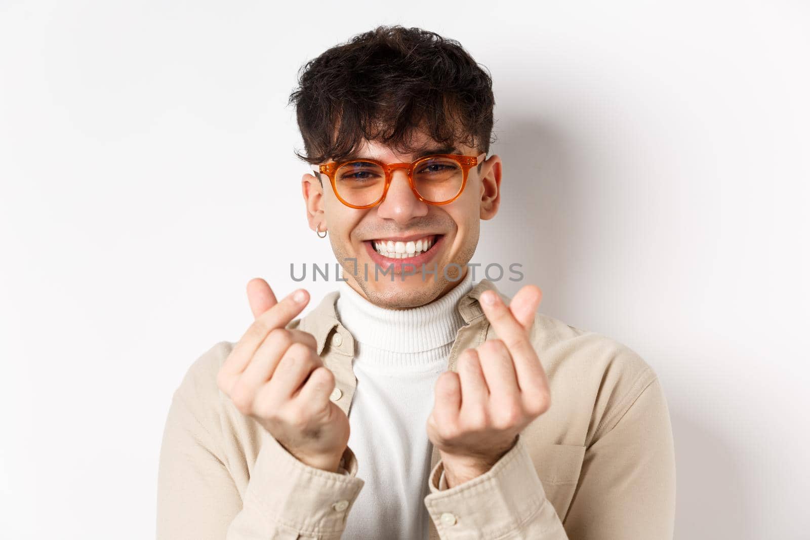 Cute young guy in glasses smiling and showing finger hearts, standing on white background.