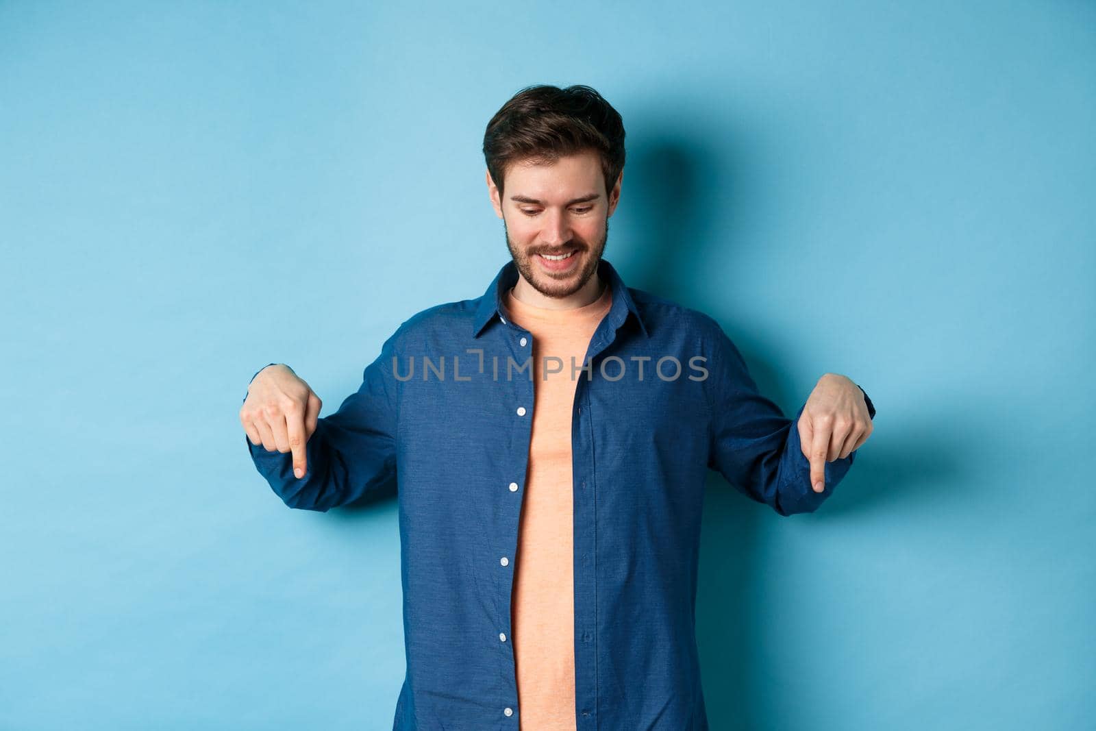 Smiling handsome man with beard, looking and pointing down at banner, checking out special offer, standing on blue background.