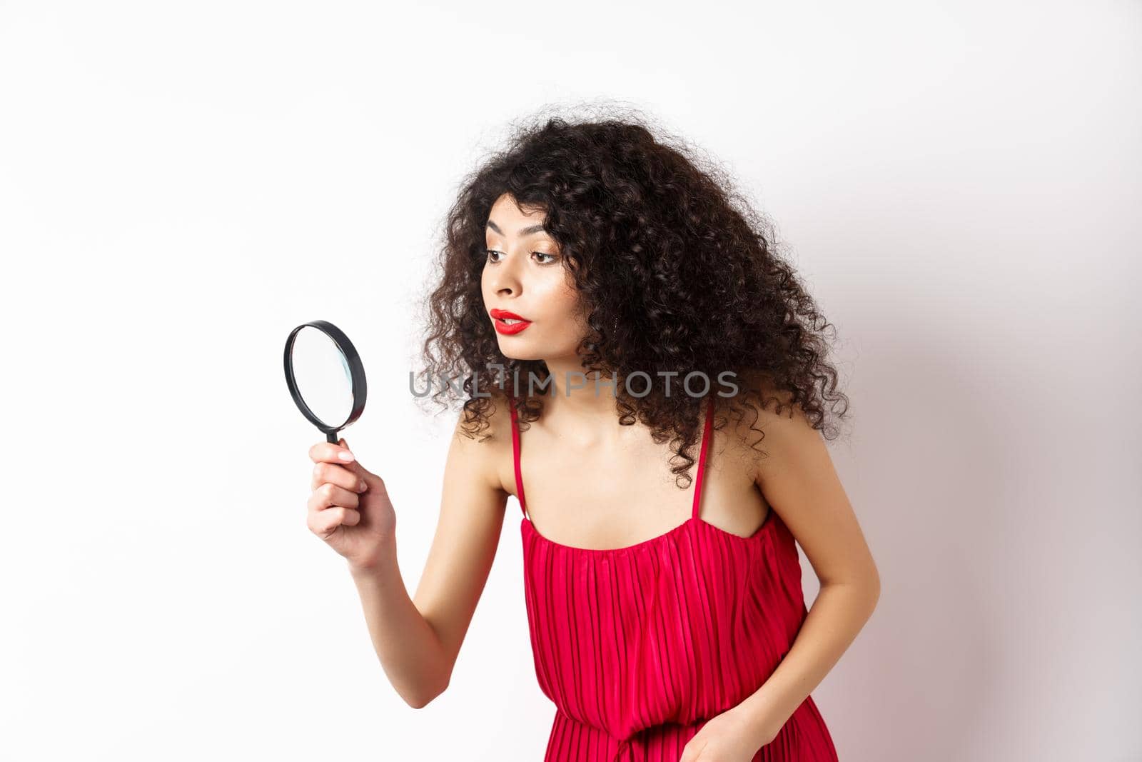 Beautiful woman in red dress searching for something, looking aside through magnifying glass, standing against white background.