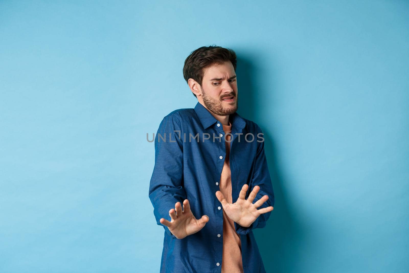 Image of man cringe and look at something disgusting, step back from awful thing and block it with hands, standing on blue background by Benzoix