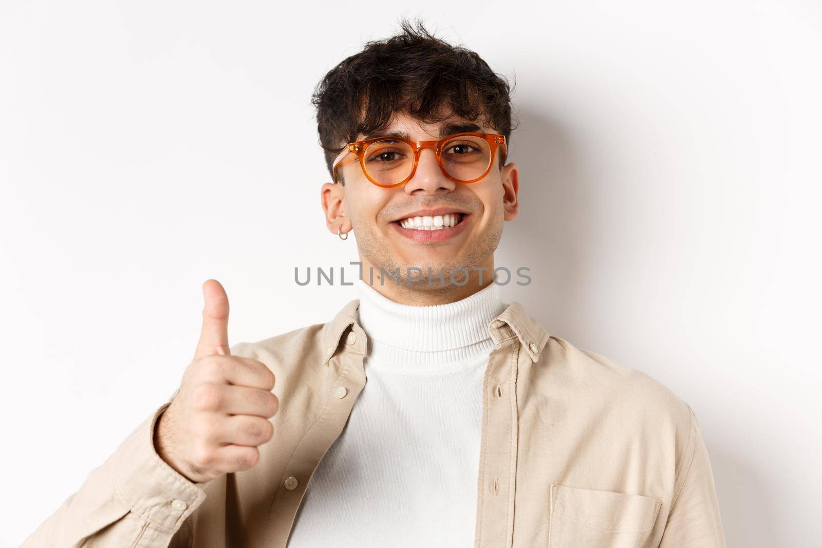 Handsome modern guy in glasses and earring, smiling satisfied and show thumb up, recommend company, standing on white background.