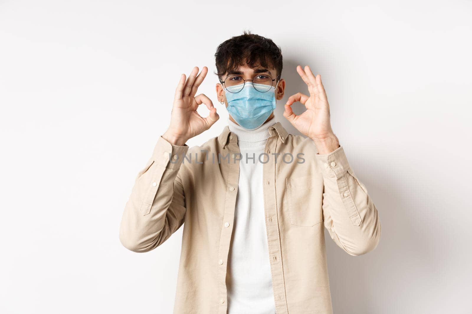 Covid-19, health and real people concept. Handsome guy in glasses and medical mask showing okay gesture in approval, recommend use preventive measures from corona, white background.