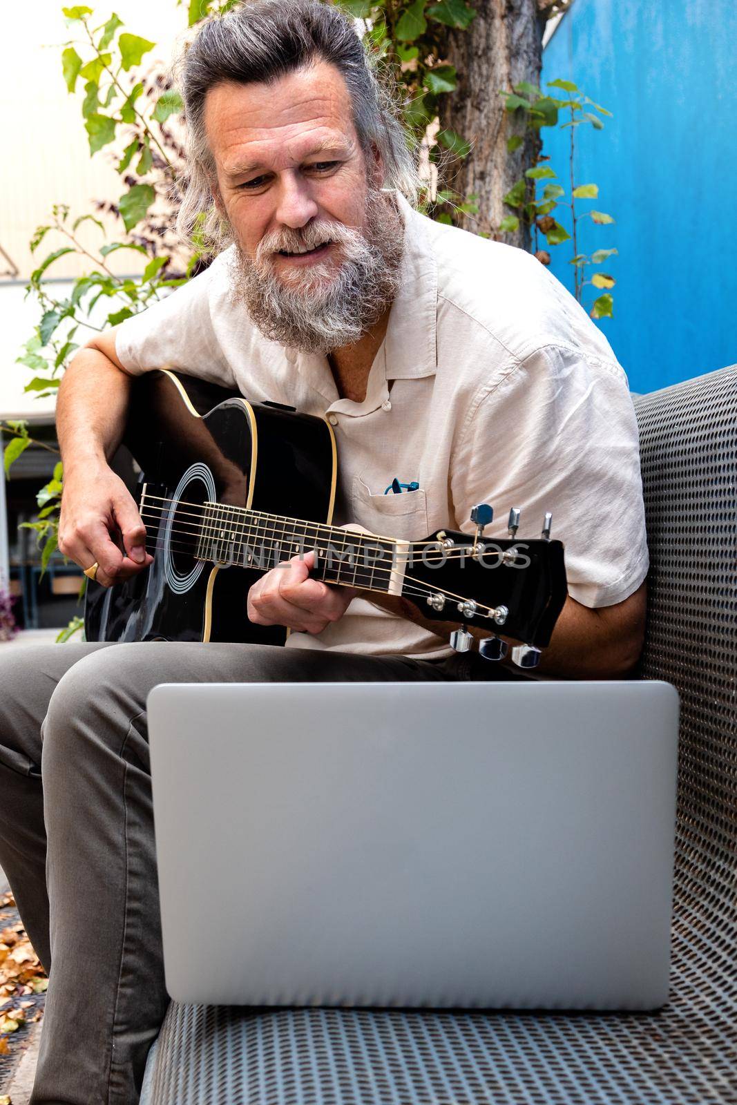 Mature caucasian man taking online guitar lessons using laptop sitting on a bench. Vertical image. by Hoverstock