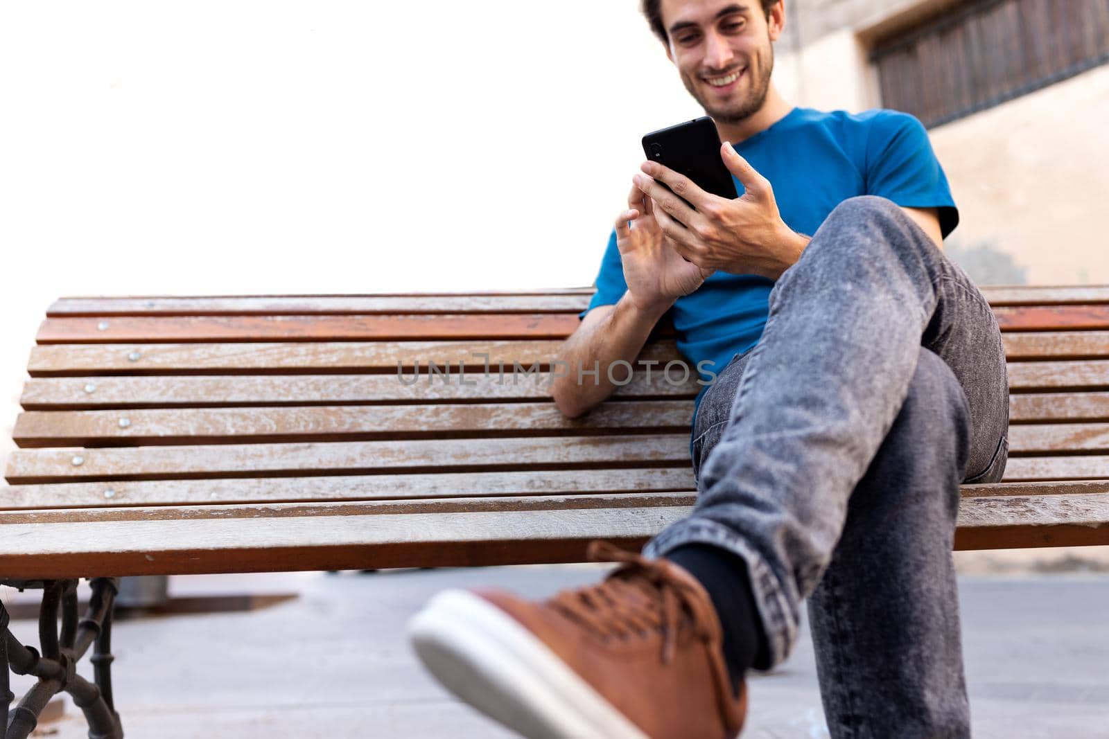 Young happy man sitting on bench using cellphone. Selective focus. Copy space. by Hoverstock