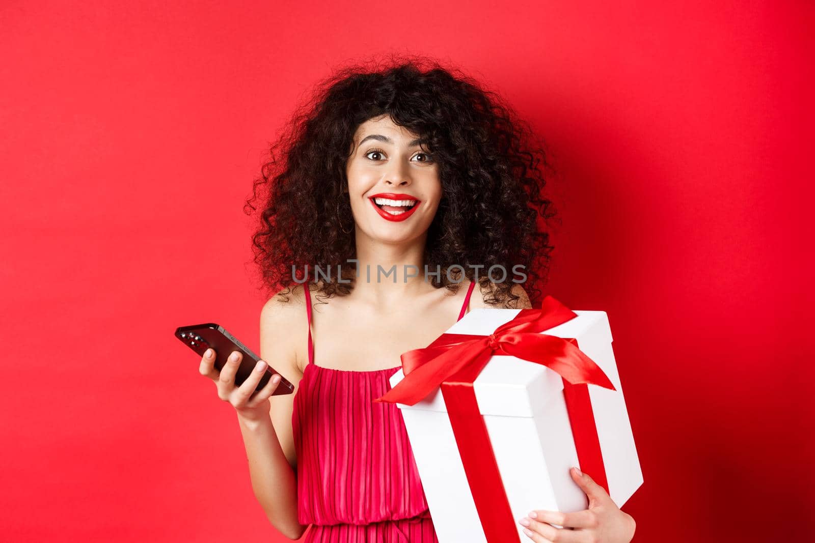 Online shopping and Valentines day. Beautiful young woman holding smartphone and lovers gift, looking surprised and happy at camera, red background.