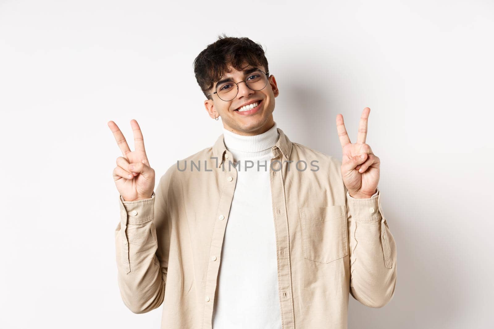 Handsome hipster guy in glasses, smiling with white teeth, showing peace or victory gesture, standing upbeat on white background.