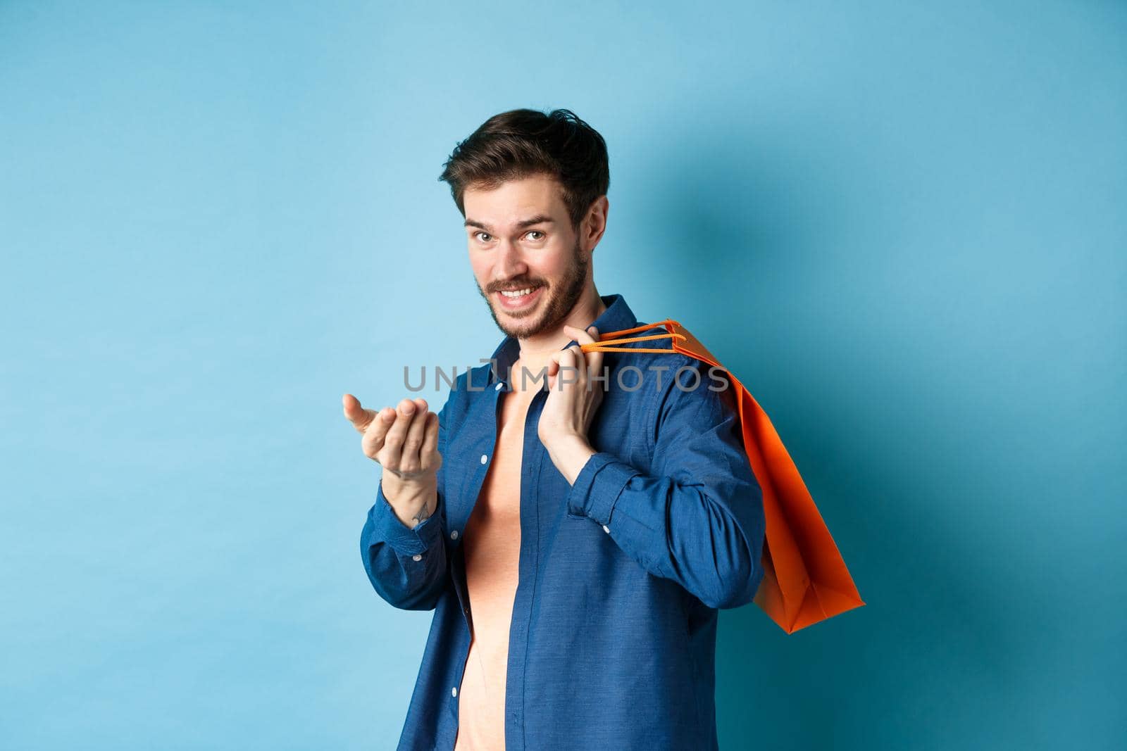Modern guy holding shopping bag behind shoulder and beckon you, telling to come closer or inviting to store, standing on blue background.