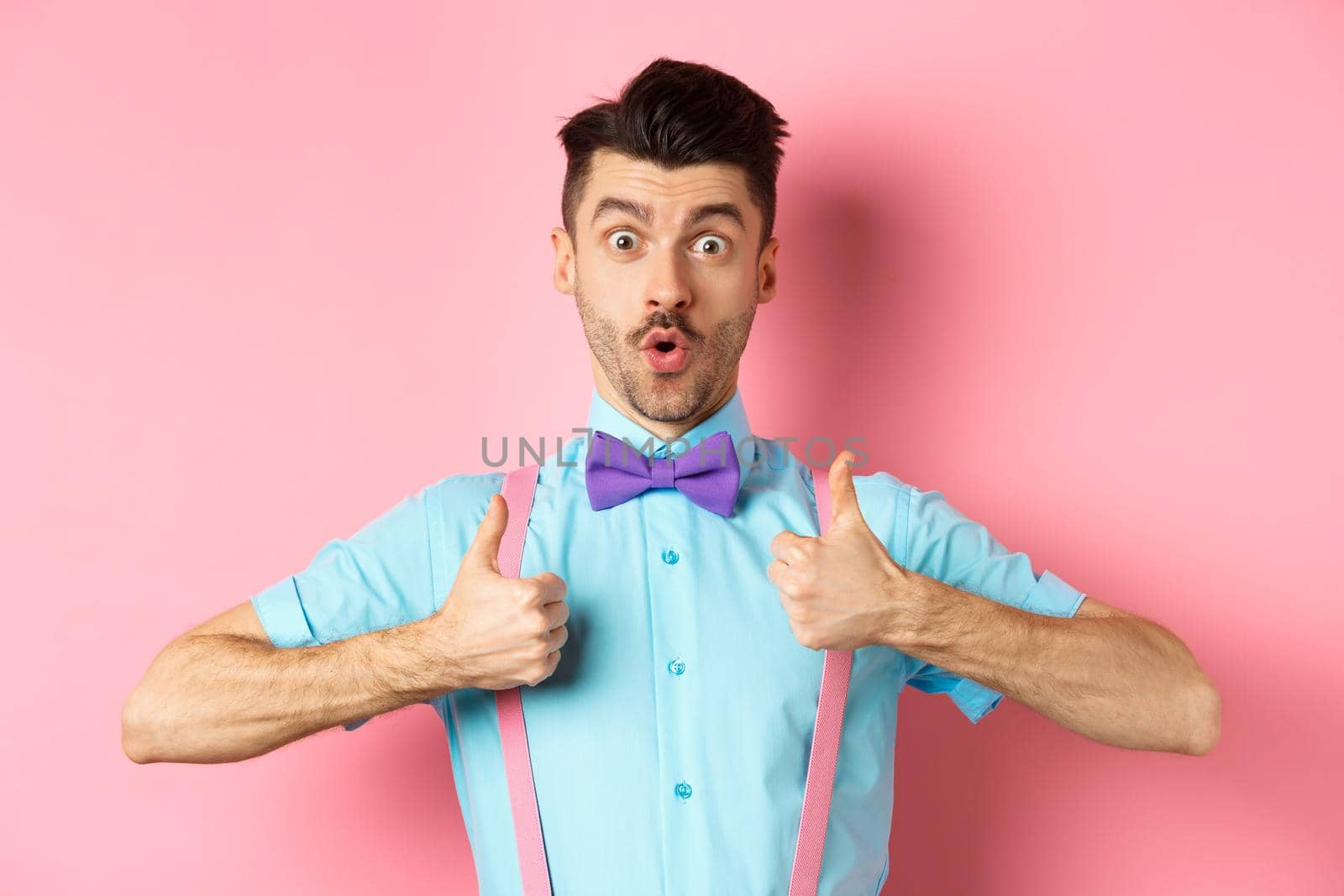 Impressed young guy in suspenders and bow-tie showing thumbs up and saying wow, recommending awesome product, praising quality, standing over pink background by Benzoix