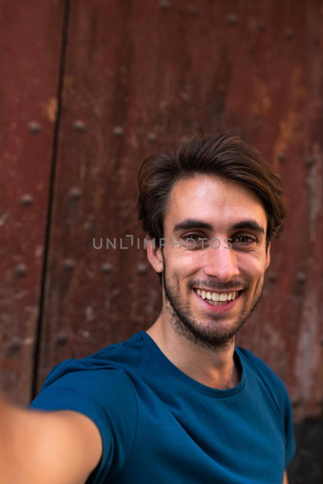 Smiling young man looking at camera takes selfie with wood background. Vertical. by Hoverstock