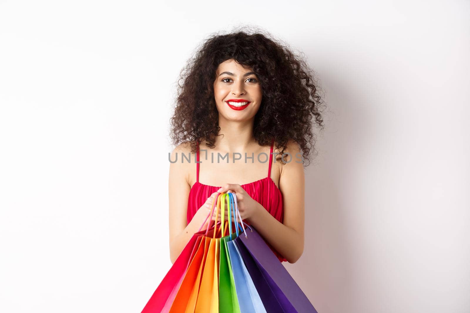 Image of stylish caucasian woman in red dress and makeup, holding shopping bags and smiling, standing over white background.