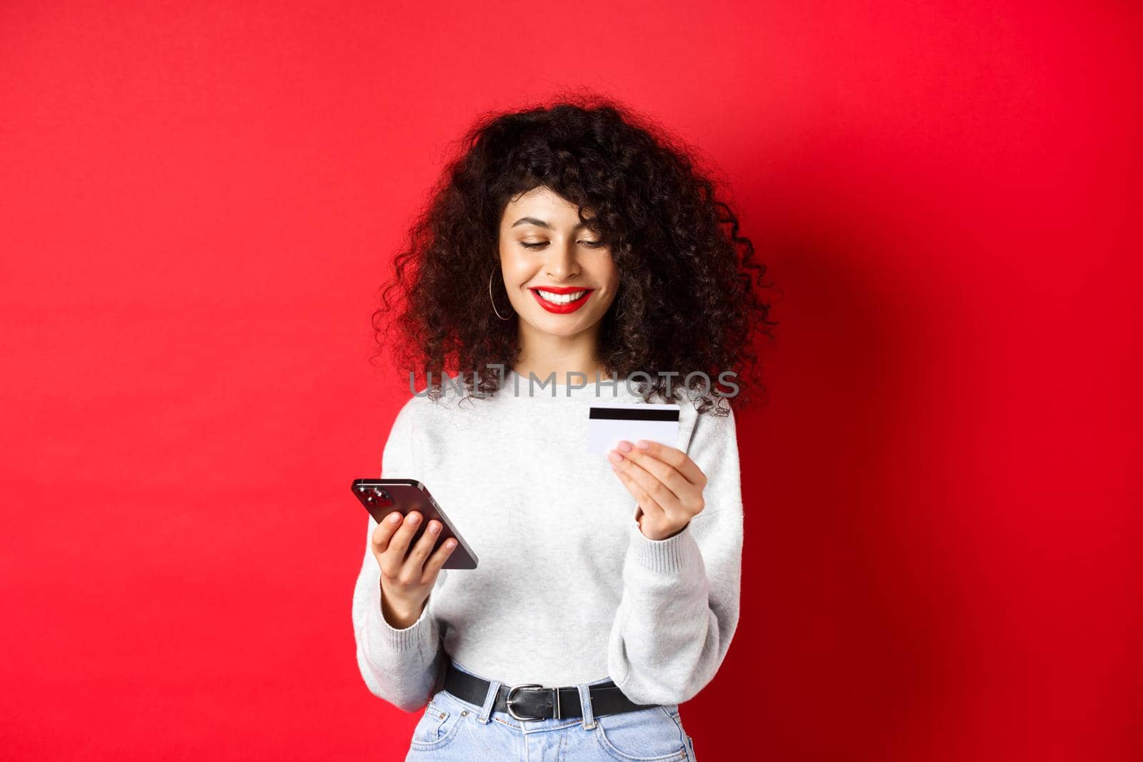 E-commerce and online shopping concept. Young modern woman paying with credit card, making purchase with smartphone, standing on red background.