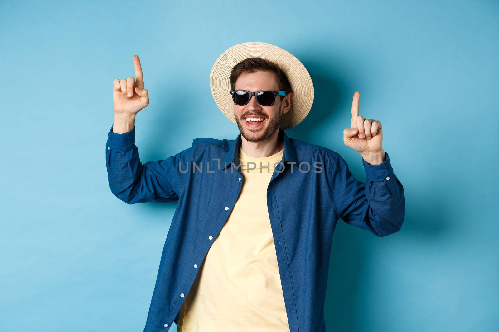Cheerful caucasian guy in straw hat and sunglasses, dancing and having fun on vacation, standing over blue background. Concept of summer tourism.