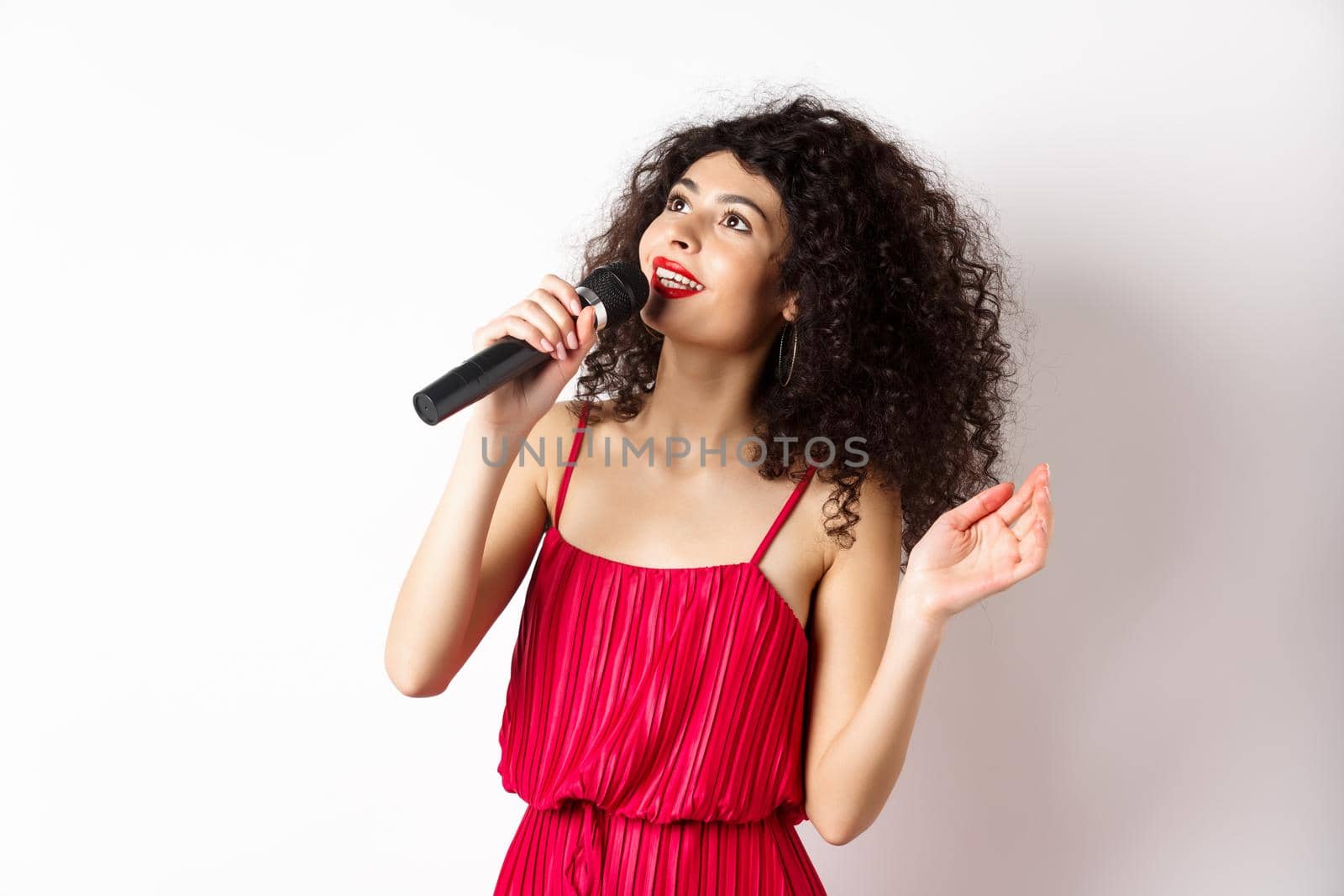 Beautiful lady in red dress singing songs in microphone, smiling and looking up, standing on white background.
